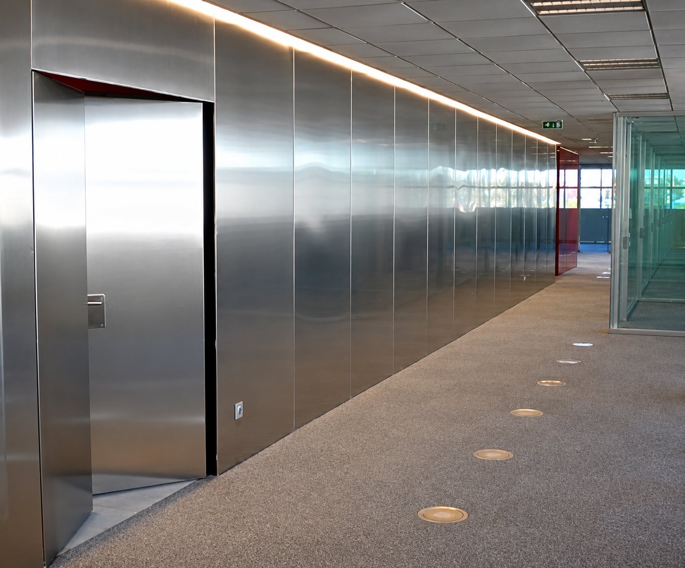 Un long couloir de bureaux dont les murs sont revêtus de panneaux d'acier inoxydable brillant, avec une porte ouverte, une moquette grise et une bande lumineuse au plafond.
