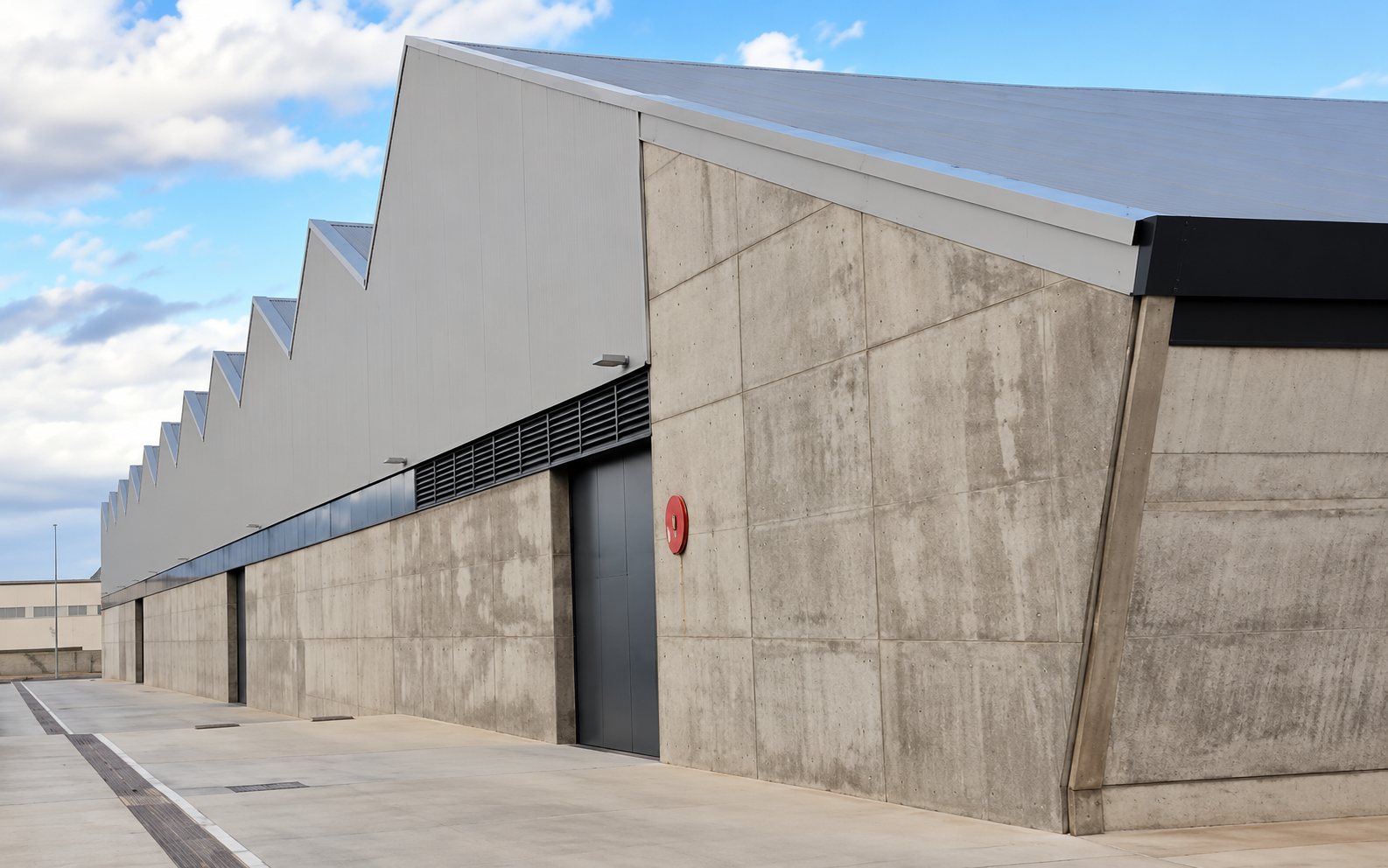 Façade d'un grand entrepôt en béton gris avec une porte métallique et une toiture en dents de scie sous un ciel nuageux.
