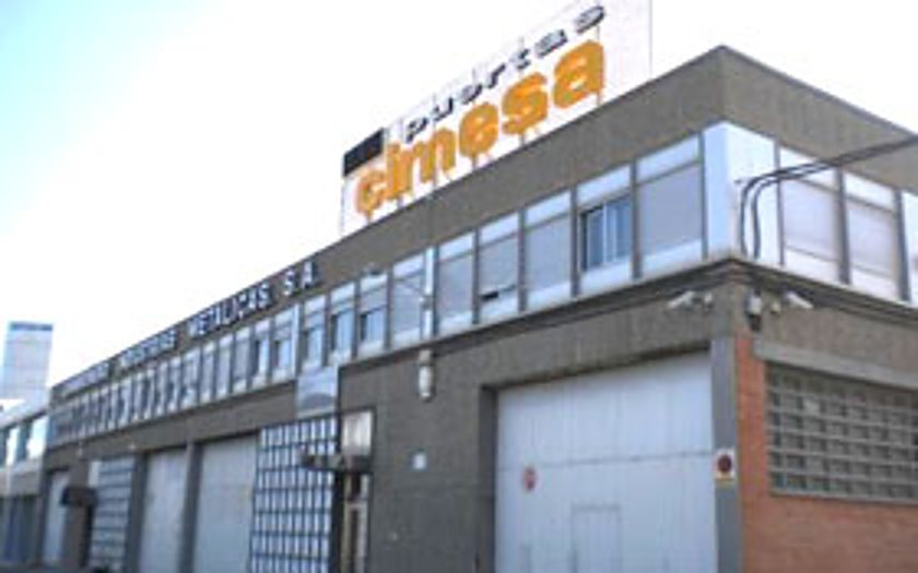 Façade d'un bâtiment industriel à deux niveaux surmonté d'une grande enseigne indiquant Puertas Cimesa, avec des portes métalliques au rez-de-chaussée.