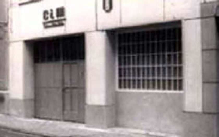 Photographie en noir et blanc de la façade d'un local commercial avec une grande porte en bois, une fenêtre à barreaux et une enseigne au-dessus.
