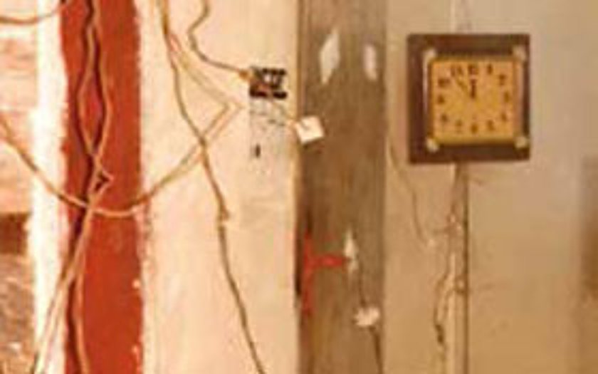 Une petite horloge carrée à cadre sombre est accrochée à un mur abîmé où pendent plusieurs fils électriques, à côté d'une bande rouge.