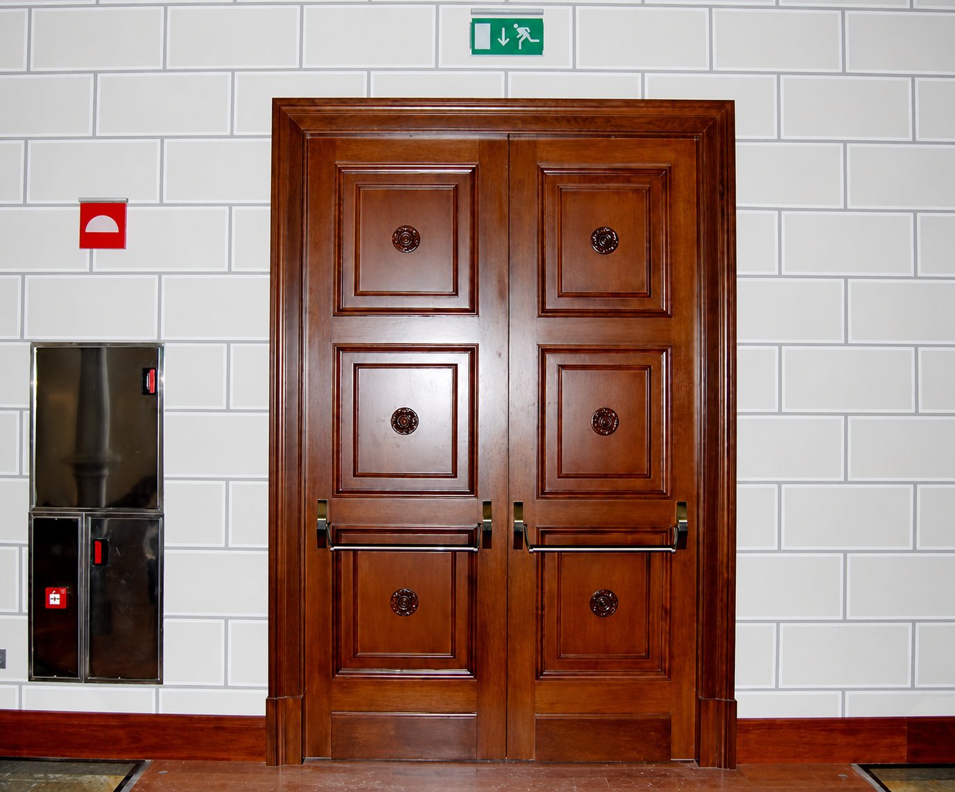Une porte à deux battants en bois foncé, ornée de panneaux moulurés et de poignées métalliques, est encadrée par un mur de pierre claire avec un panneau de sortie de secours vert au-dessus.