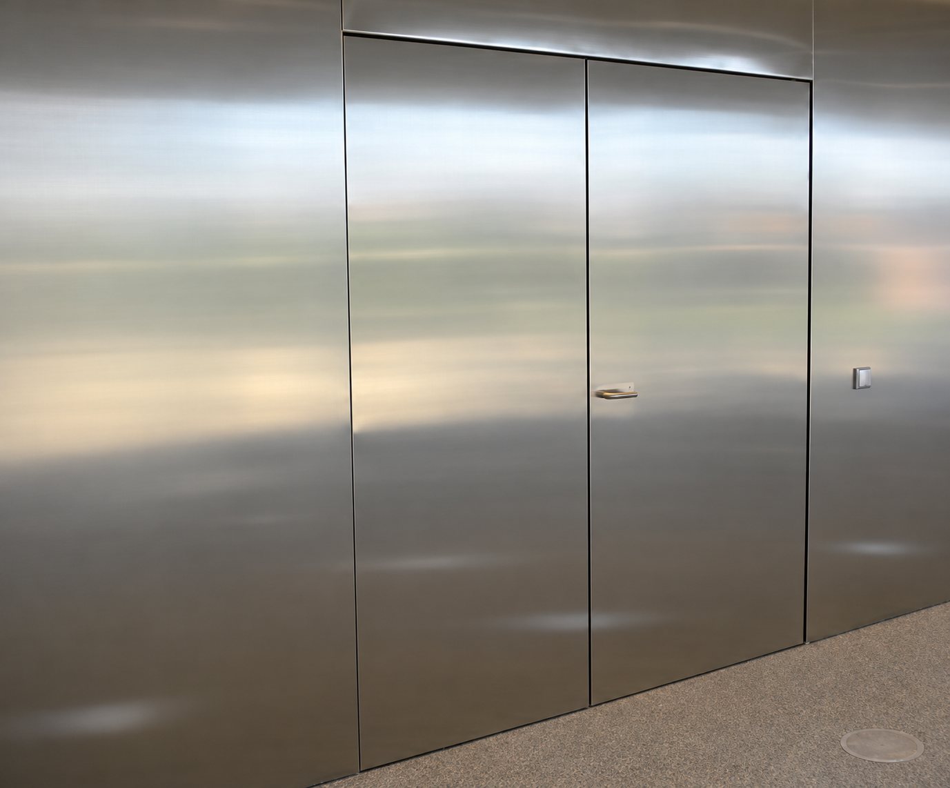 Une porte double en acier inoxydable brossé, affleurante, est intégrée dans un mur entièrement revêtu de métal, avec une poignée discrète et un moquette grise au sol.