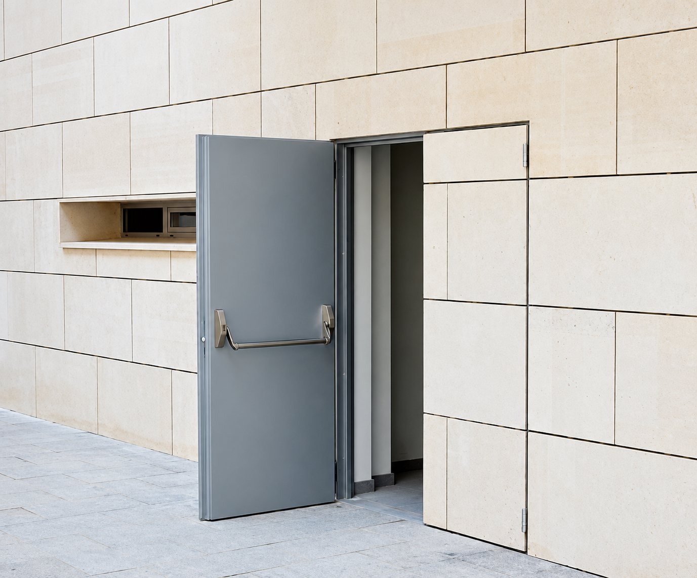 Une porte coupe-feu grise avec barre antipanique est entrouverte dans un mur de façade en dalles de pierre beige, à côté d'une niche technique.