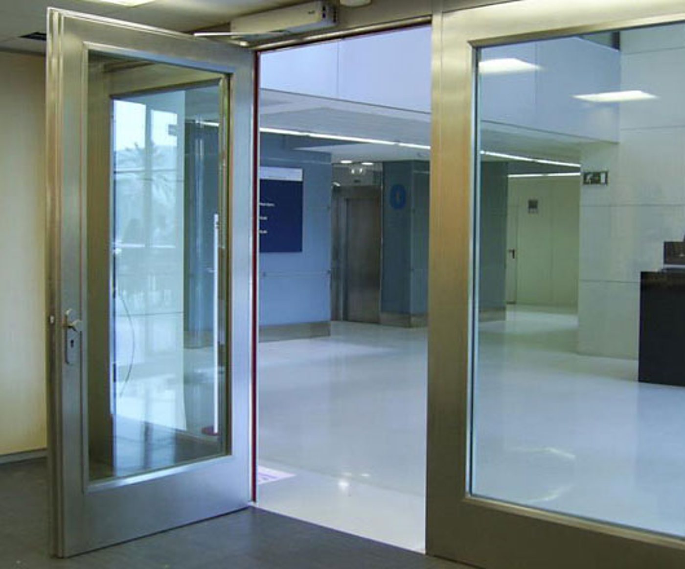 Une porte vitrée à cadre en acier inoxydable est ouverte et laisse voir un grand hall moderne avec un sol brillant et des panneaux bleus.