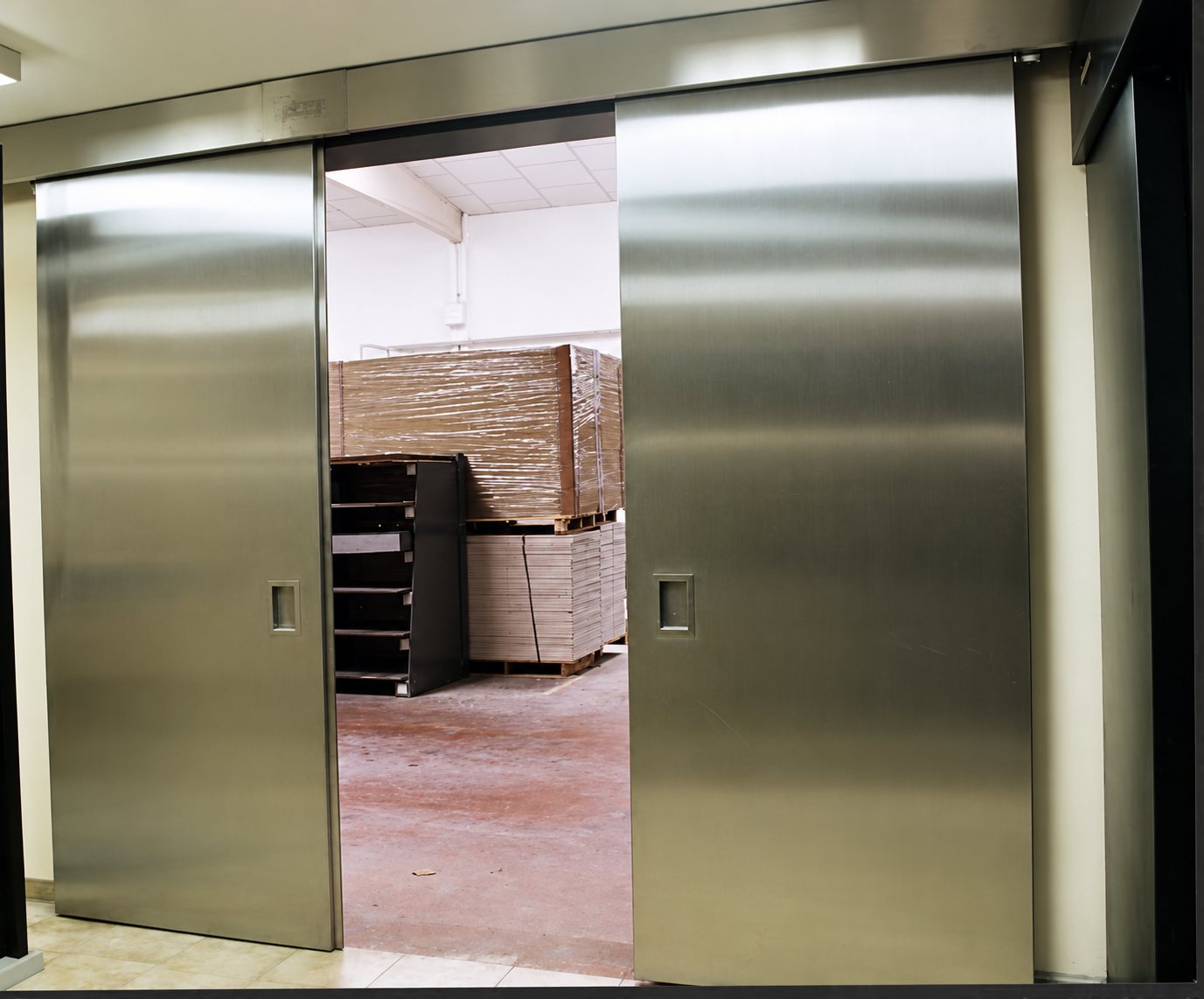 Deux grandes portes coulissantes en acier inoxydable entrouvertes laissent voir des palettes de cartons empilées dans un entrepôt.